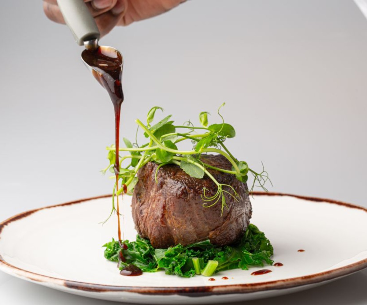 Fillet steak on a white plate sat on kale and garnished with pea shoots, drizzled with a sauce